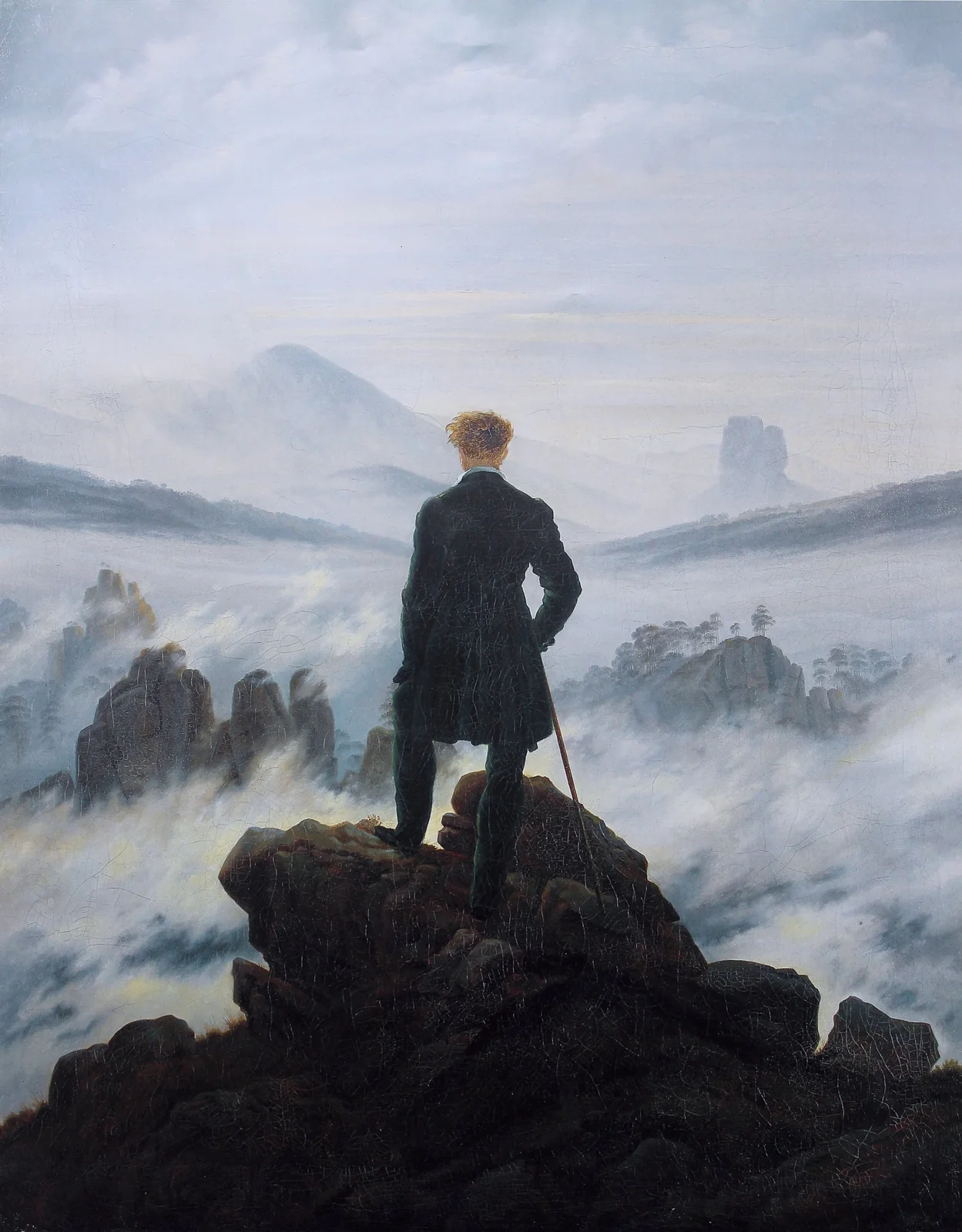 A figure with a walking stick stands on a rocky summit, facing mountains partly hidden by fog.